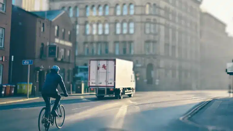 Cyclist riding near an HGV on a city street with clear road markings and historic buildings in the background.